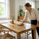Person gently wiping down a modern wooden dining table with a yellow microfiber cloth in a sunlit room