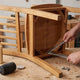 Person tightening corner bracket bolts under a wobbly wooden chair on a workbench with wood glue and mallet nearby